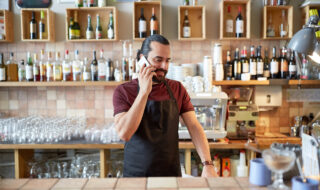 small business, communication, people and service concept - happy man or waiter in apron calling on smartphone at bar or coffee shop