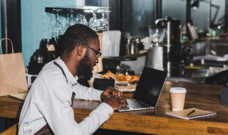 smiling young african american barista in eyeglasses using laptop in coffee shop