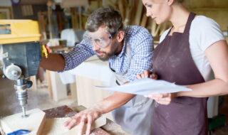 Portrait of two carpenters, man and woman, operating drilling machine making furniture in woodworking shop
