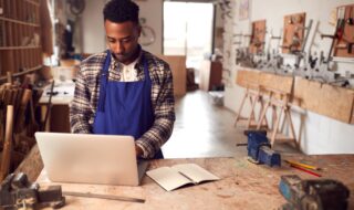 African American on a computer while at his wood workshop wearing a blue smock
