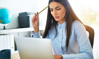 Young Asian businesswoman frowning with concern as she tries to understand something she is reading on her laptop computer scratching her head with her pencil in perplexity