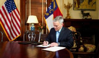 North Carolina Gov. Josh Stein sitting at a desk in front of the state flag, signing executive orders providing relief for people affected by Hurricane Helene