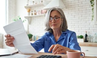 Woman sitting down at a kitchen table with a piece of paper in her left hand while using a calculator