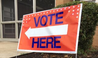 Large Orange background colored placard with Bold blue lettering that says Vote here