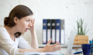 Young stressed businesswoman sitting with laptop and touching head with shocked facial expression. Surprised business person looking at laptop computer worried and amazed with open mouth and big eyes