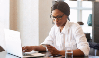 Young woman with glasses looking at a credit card while using a computer during the daytime indoors